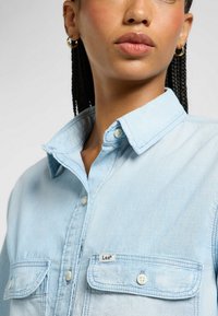 Camisa de mezclilla de color azul claro con diseño de botones, dos bolsillos frontales y cuello. La tela tiene una textura suave.