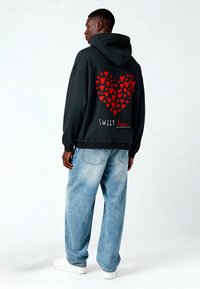 Felpa con cappuccio nera con una grafica a forma di cuore rosso composta da cuori più piccoli, con il testo "SWEET heart" sotto. Indossata con jeans larghi di colore azzurro chiaro.