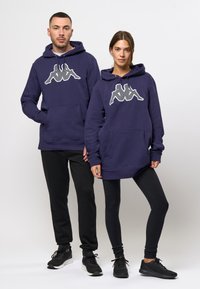 Sudaderas con capucha en color navy con un gran logotipo blanco, combinadas con leggins negros y zapatillas deportivas. Presentan un bolsillo frontal y puños acanalados.
