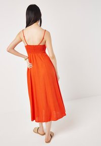 Rotes, ärmelloses Maxikleid mit gesmoktem Oberteil, fließendem Rock und verstellbaren Spaghettiträgern. Lässiger Stoff; getragen mit flachen Sandalen.