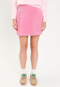 Kobieta w różowej mini spódnicy z korduroju, beżowych sneakersach z zielonymi sznurowadłami, białych skarpetach i jasnoróżowym swetrze na białym tle.