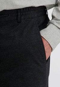 Hand in de zak van donkere, gestructureerde broek in combinatie met een lichtgrijs langmouwshirt, met de manchet zichtbaar tegen een effen achtergrond.
