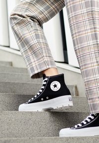 Czarne płócienne trampki wysokie z białą gumową podeszwą, z okrągłą naszywką logo i białymi sznurówkami, noszone z spodniami w kratę na schodach.