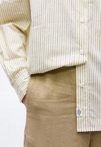 Μπεζ και λευκό ριγέ πουκάμισο με κουμπιά, φορεμένο μέσα σε ανοιχτό καφέ παντελόνι, με το ένα χέρι εν μέρει στην τσέπη.