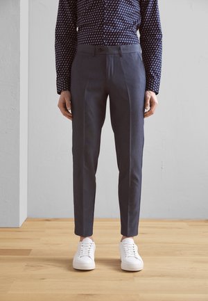 Dunkelblaue, maßgeschneiderte Hose mit glatter Textur und Vorderfalten, kombiniert mit weißen Sneakers, stehend auf einem Holzboden.
