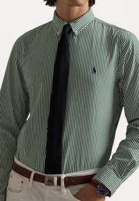 Grün-weiß gestreiftes Hemd mit Knöpfen und einer marineblauen Krawatte. Es verfügt über ein kleines Logo auf der Brust, kombiniert mit einem braunen Gürtel und weißen Hosen.