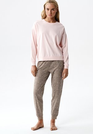 Młoda kobieta w jasnoróżowej bluzce z długim rękawem i beżowych spodniach dresowych w cętki lamparta, stoi boso na białym tle.