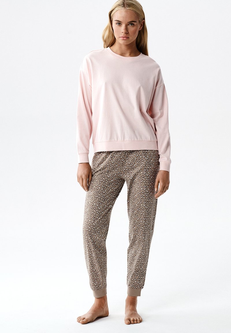 Młoda kobieta w jasnoróżowej bluzce z długim rękawem i beżowych spodniach dresowych w cętki lamparta, stoi boso na białym tle.