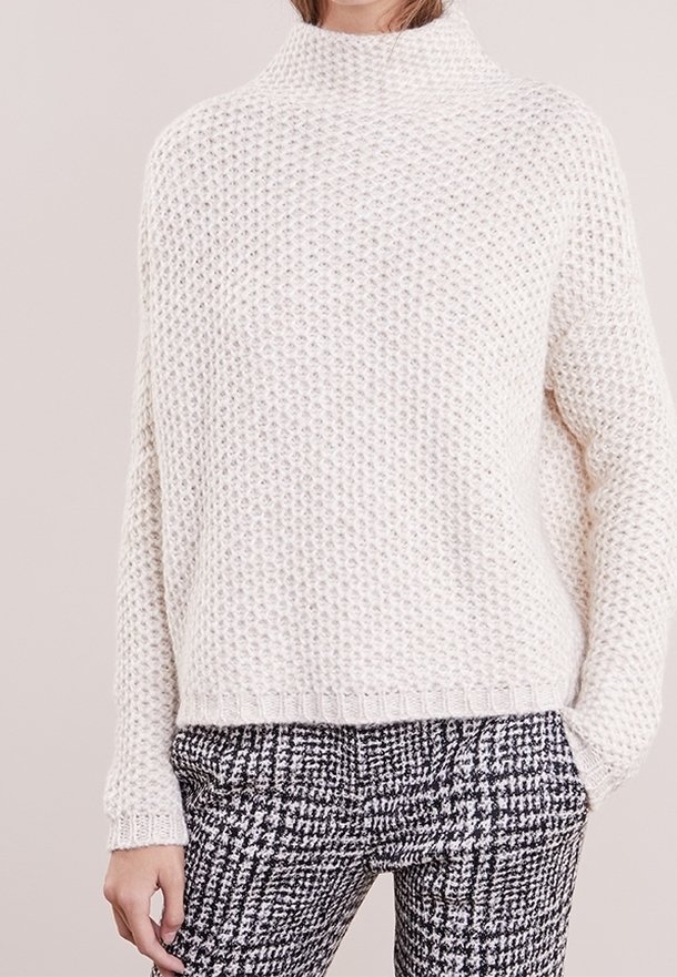 Pull épais en tricot couleur crème, à col montant, manches longues et motif texturé, associé à un pantalon à motif pied-de-poule noir et blanc.