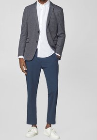 Blazer grigio a pattern sopra una camicia bianca con colletto, abbinato a pantaloni blu navy e sneakers bianche. Linee pulite, vestibilità su misura, look moderno.