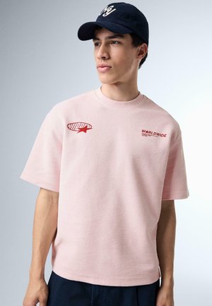 Mladý muž nosi svetloružovú krátky rukáv košeľu s červenými logami a tmavomodrú čiapku, pozerá sa do strany na neutrálnom pozadí.