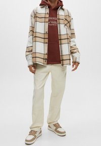 Chemise en flanelle blanche à carreaux marron, portée sur un sweat à capuche bordeaux. Un pantalon clair et des baskets beige complètent la tenue.