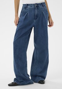 Laiade jalaga, kõrge vöökohaga teksad tumedas sinises denimis. Omadusteks ees voldid, sile tekstuur ja klassikaline viie taskuga disain koos nööpidega kinnitusega.