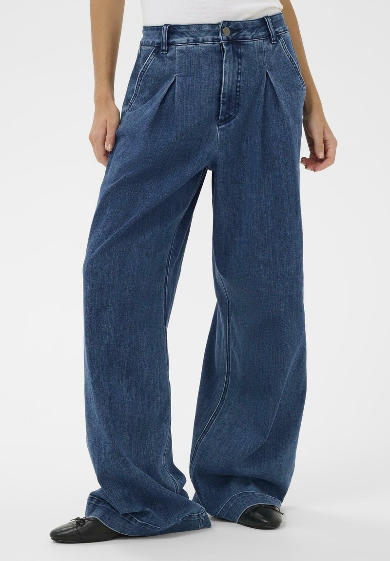 Laiade jalaga, kõrge vöökohaga teksad tumedas sinises denimis. Omadusteks ees voldid, sile tekstuur ja klassikaline viie taskuga disain koos nööpidega kinnitusega.