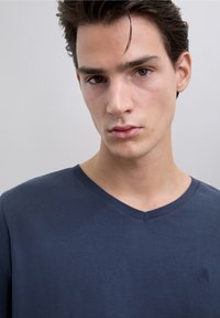 Camiseta azul marino con cuello en V, hecha de tela suave, con costuras mínimas y un pequeño logo en el pecho. Textura lisa, diseño casual.