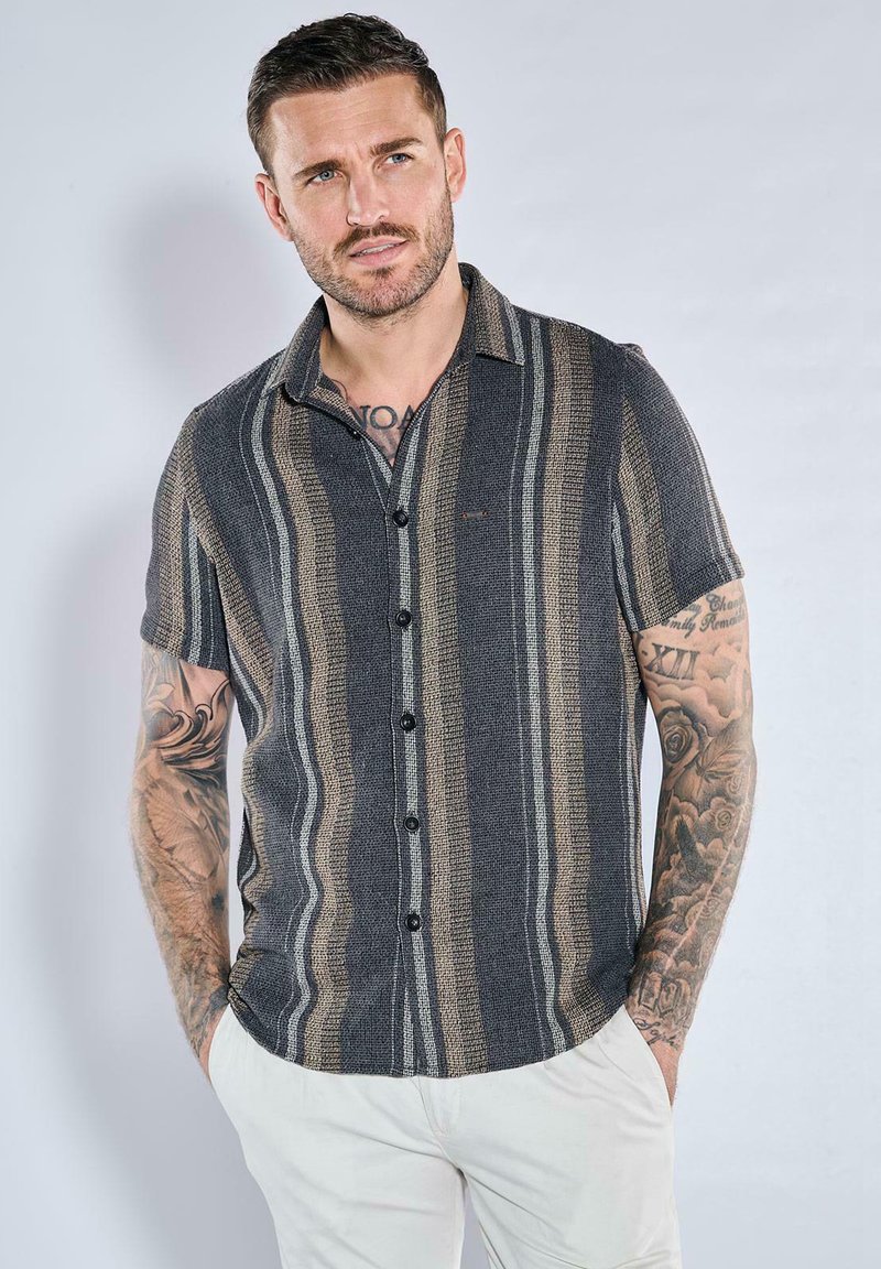 Man with tattoos wearing a short-sleeve button-up shirt with vertical stripes and white pants, standing with hands in pockets against light background.