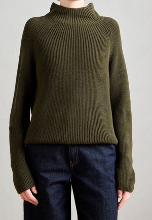 Personne portant un pull en maille côtelée vert olive à manches longues et col montant, associé à un jean bleu foncé sur un fond uni.