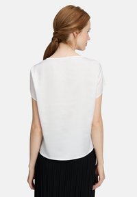 Weiße kurzärmelige Bluse aus glattem Stoff, mit einem runden Ausschnitt und einer lockeren Passform. Der Rücken ist etwas länger und hat einen abgerundeten Saum.