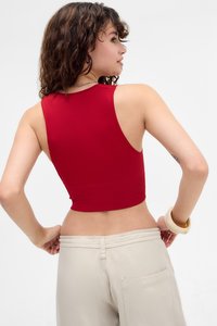 Žena s kovrčavom kosom koja nosi bezrukavnu crvenu majicu i bež hlače, okrenuta blago da pokaže leđa i bočni profil.