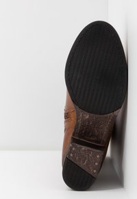 Botte en cuir marron avec une semelle en caoutchouc noire présentant un motif de crampons en zigzag, positionnée verticalement contre un mur blanc.