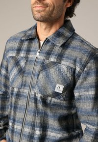 Geruit flanellen shirt in blauw, grijs en zwart. Kenmerken zijn een volledige rits, twee borstzakken en een labelaccent, met een textuurstof.