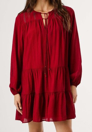 Femme portant une robe rouge ample à manches longues à étages, avec un décolleté en trou de serrure noué par de fines cordelettes et de petites perles métalliques.