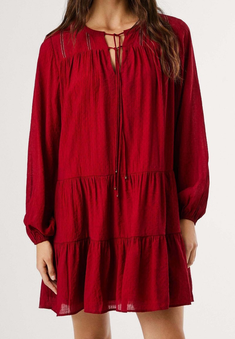 Femme portant une robe rouge ample à manches longues à étages, avec un décolleté en trou de serrure noué par de fines cordelettes et de petites perles métalliques.