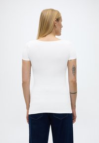 Plavuša okrenuta leđima, nosi bijelu usku majicu kratkih rukava i tamnoplave traperice, s nekoliko tetovaža na rukama.