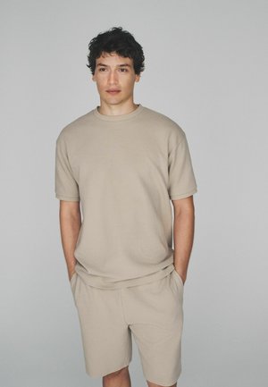 Tumejuuste ja tumedate lokkidega mees kannab beežikat lühikeste varrukatega särki ja sobivaid lühikesi pükse, seisab käed taskutes lihtsa halli tausta ees.