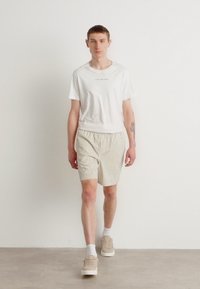 Camiseta blanca con logo, pantalones cortos rayados beige, calcetines blancos y zapatillas beige. Modelo de pie contra un fondo blanco liso.