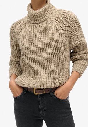 Beige chunky knit turtleneck sweater with rolled sleeves tucked into high-waisted black jeans with brown belt, hands in pockets.