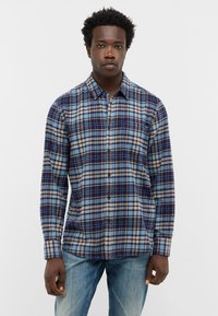 Jonge man met kort, krullend haar, gekleed in een blauw en beige geruit overhemd met knopen en vervaagde blauwe jeans, staand tegen een witte achtergrond.