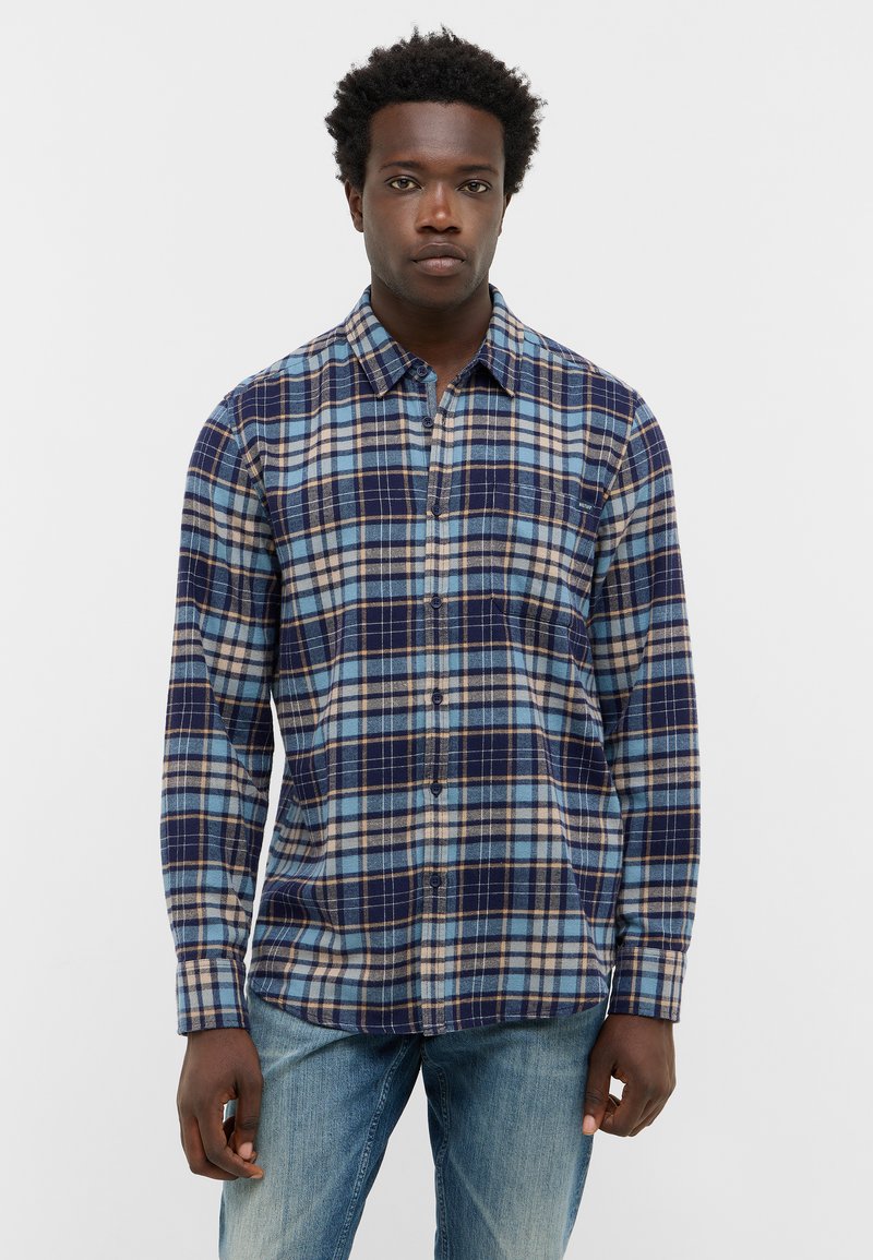 Jonge man met kort, krullend haar, gekleed in een blauw en beige geruit overhemd met knopen en vervaagde blauwe jeans, staand tegen een witte achtergrond.