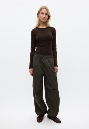 Blonde Frau trägt ein eng anliegendes braunes Langarmshirt, lockere dunkelgrüne Cargohosen und dunkelbraune Schuhe, stehend vor einem weißen Hintergrund.
