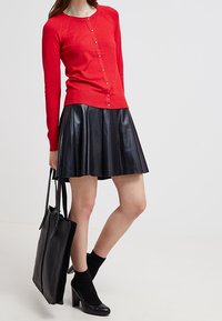 Woman wearing a red buttoned cardigan, black pleated leather skirt, black heeled shoes with socks, holding a large black tote bag.