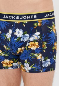 Jūras zils ziedu bokseršortus ar dzeltenu elastīgu jostu no Jack & Jones. Iezīmes zili un zeltaini ziedi, kā arī zaļi lapu raksti. Stiepļu audums.
