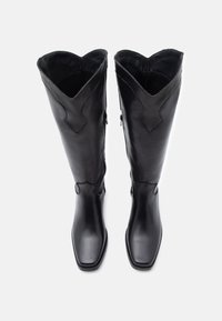 Μαύρα δερμάτινα μπότες μέχρι το γόνατο με τετράγωνη μύτη, με κυματιστό άνω άκρο και μικρά μεταλλικά διακοσμητικά στον αστράγαλο. Λεπτή υφή.