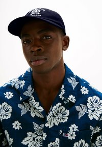 Granatowa czapka z białym logo, noszona z niebieską koszulą w kwiatowy wzór z białymi kwiatami i zielonymi liśćmi. Koszula ma kołnierzyk i kieszeń.