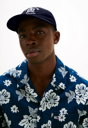 Marinepet met wit logo, gedragen met een blauw bloempatronen shirt met witte bloemen en groene bladeren. Het shirt heeft een opstaande kraag en een zak.