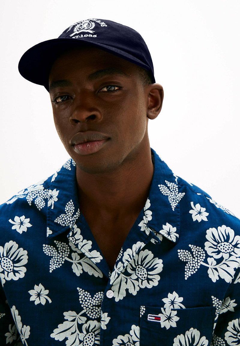 Granatowa czapka z białym logo, noszona z niebieską koszulą w kwiatowy wzór z białymi kwiatami i zielonymi liśćmi. Koszula ma kołnierzyk i kieszeń.