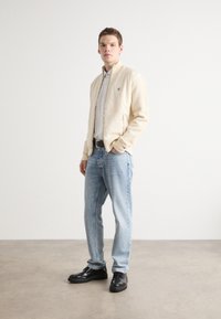 Ljus beige bomberjacka med dragkedja, bärs över en skjorta med knappar; matchas med ljusblå jeans och svarta skor. Enkelt design och rena linjer.