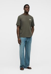 Άνδρας στέκεται φορώντας μια ελιά πράσινη μπλούζα με κείμενο, ανοιχτό μπλε φαρδιά τζιν παντελόνια και σκούρα καφέ σανδάλια μπροστά σε απλό λευκό φόντο.