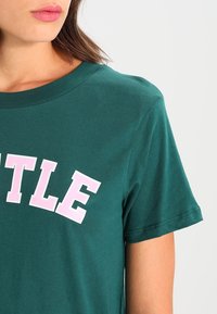 Teal T-shirt med korte ærmer, rund halsudskæring og fed pink skrift, der står "SEATTLE." Bomuldsstof med glat overflade.