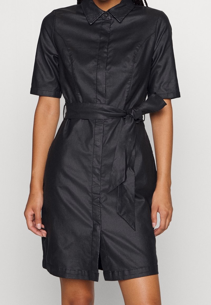 Robe chemise noire à manches courtes avec col et ceinture nouée à la taille, portée par une personne à la peau de teinte moyen-foncée.