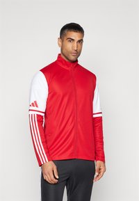 Rode sportjack met ritssluiting en witte mouwen, voorzien van drie strepen op elke arm en een hoge kraag. Glad textuur, atletische pasvorm.