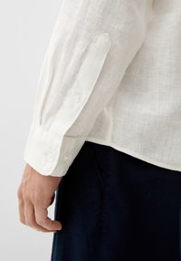 Weißes Leinenhemd mit einem geknöpften Manschette, das aus einem strukturierten, atmungsaktiven Stoff besteht. Die Manschette hat ein spitzes Design und zwei Knöpfe.