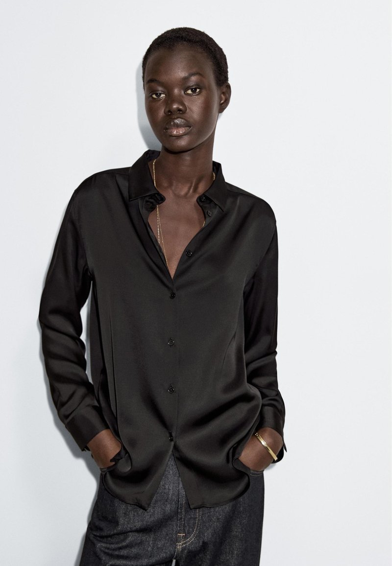 Czarna satynowa koszula na guziki z wyraźnym kołnierzykiem, długimi rękawami i luźnym krojem, połączona z ciemnymi dżinsami.