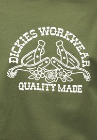 Πράσινο ύφασμα με λευκό κείμενο "Dickies Workwear Quality Made" και γραφικό δύο σταυρωτών καουμπόηδων με αγκάθια και λουλούδια από κάτω.