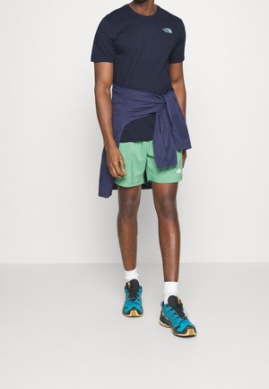 Homme portant un t-shirt bleu marine, un short vert, une veste bleue nouée autour de la taille, des chaussettes blanches et des chaussures de randonnée bleues, debout sur un fond uni.