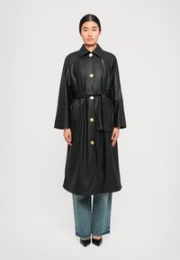 Cappotto trench in finta pelle nera con grandi bottoni dorati, vita con cintura e tasca con zip. Caratterizzato da maniche ampie e design lungo fino al ginocchio.